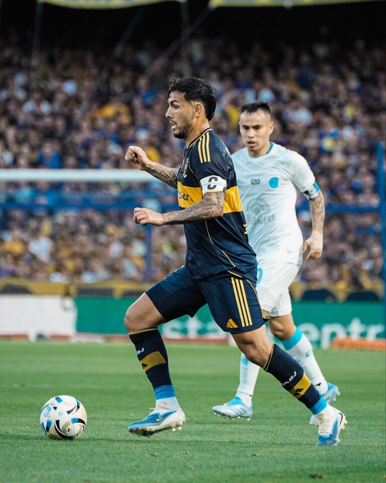 A vibrant scene from the Boca Juniors vs. Belgrano football match at La Bombonera stadium in 2025. The image captures a packed crowd of passionate Boca Juniors fans in blue and yellow, waving flags and cheering, with the iconic stadium's architecture in the background. The field shows players in action, with Belgrano in white and blue kits celebrating a goal, while Boca players in their traditional blue and gold kits react. The atmosphere is electric, with confetti and banners adding to the intensity of the Argentine football rivalry.